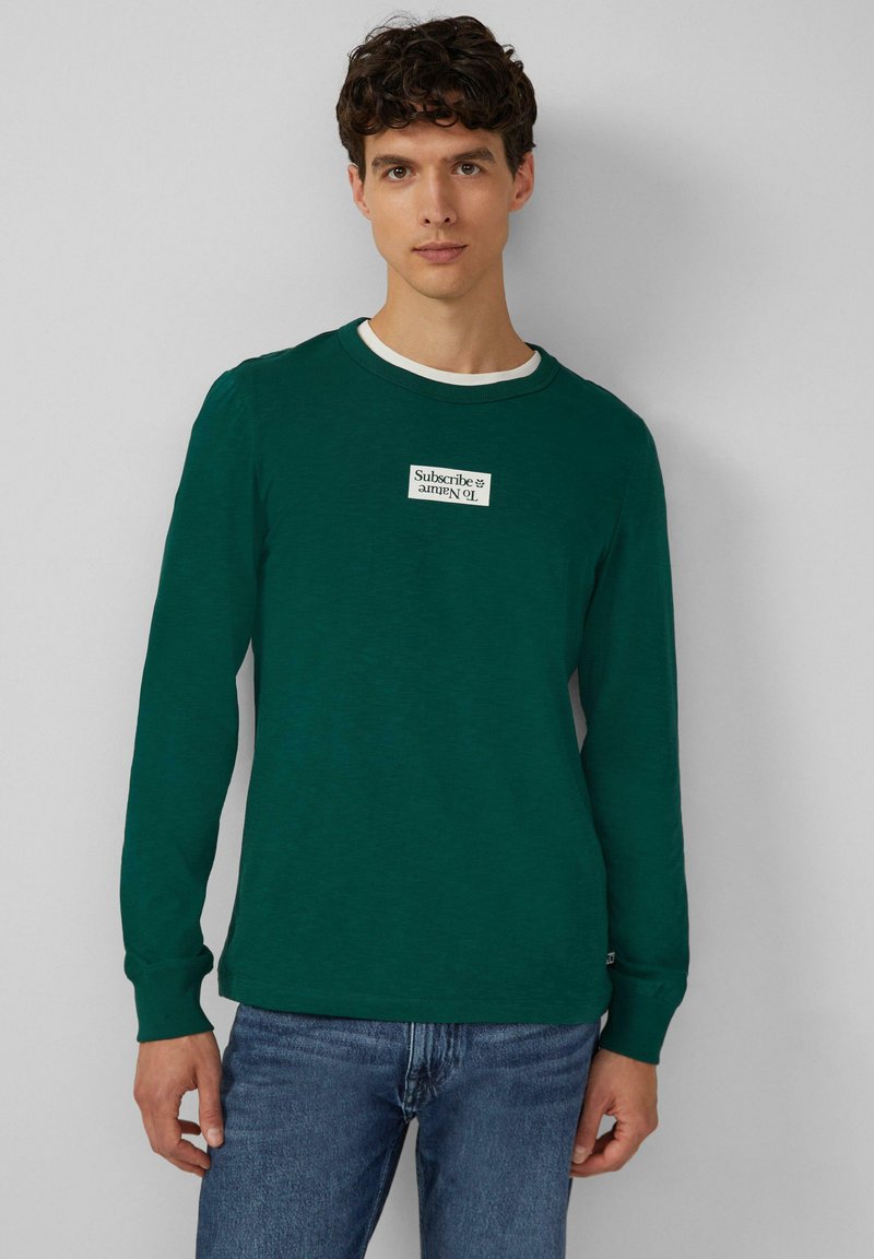 Zielona longsleeve z miękkiego materiału, z białym dekoltem i naszywką z napisem "Subskrybuj" na piersi.