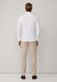 Biała, długiego rękawa koszula z zapięciem na guziki, w połączeniu z beżowymi spodniami cargo z bocznymi kieszeniami, noszona z ciemnymi butami, widok z tyłu.