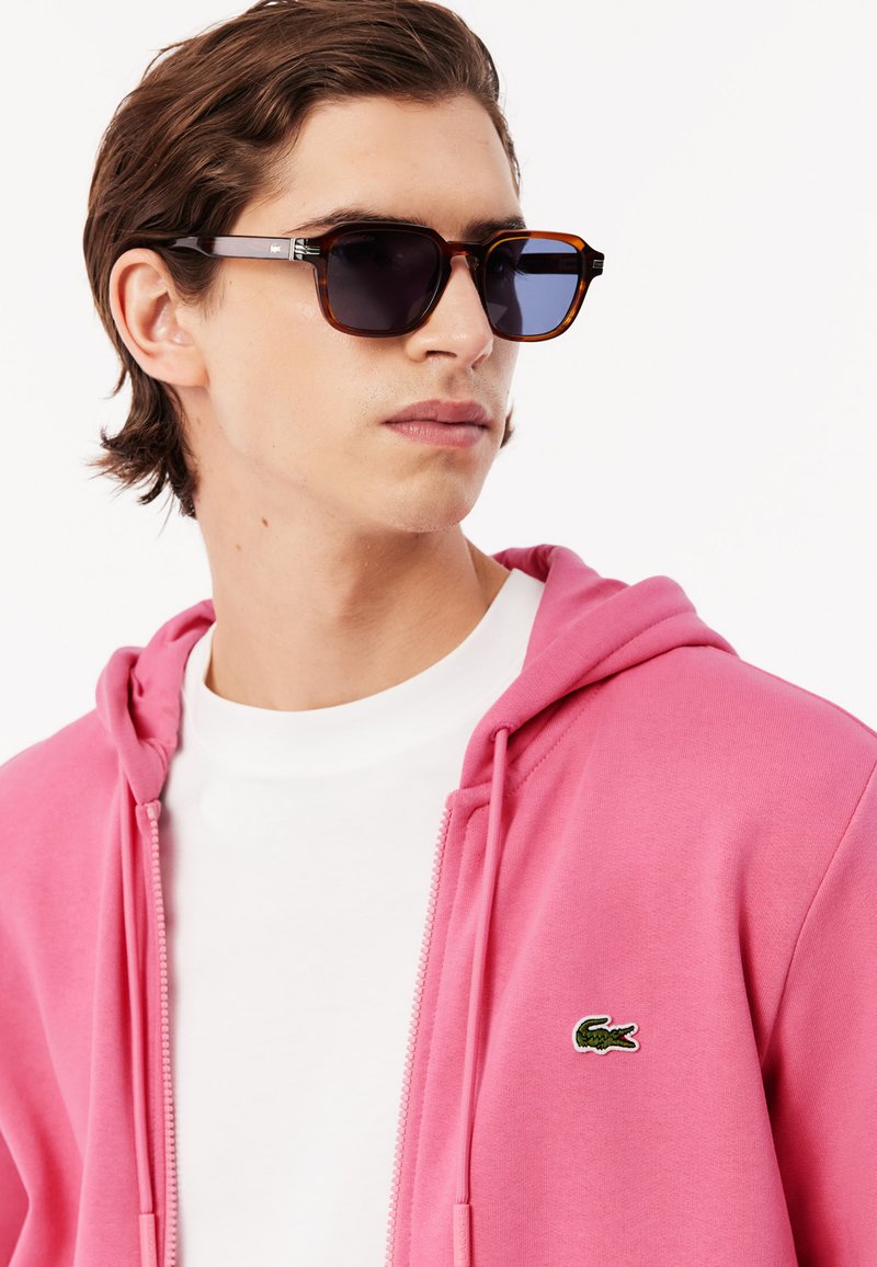 Occhiali da sole con lenti scure e montatura tartarugata. Il modello indossa un hoodie rosa e una t-shirt bianca, con un piccolo logo verde sull'hoodie.