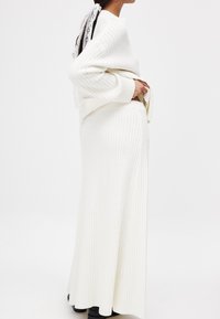 Häkelweißer Pullover und passender Maxi-Rock mit geripptem Muster. Das Modell trägt ein haaraccessoire mit Spitze und schwarze Ankle Boots.