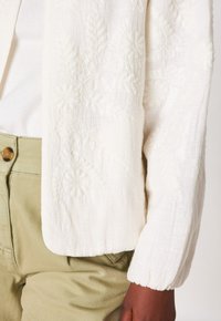 Giacca bianca a maniche lunghe con texture e ricami floreali indossata sopra un top bianco, abbinata a pantaloni verde chiaro a vita alta con chiusura a bottone.