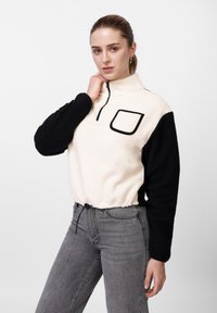 Πουλόβερ από φλις σε κρεμ και μαύρο, με ψηλό γιακά, μισό φερμουάρ και μπροστινή τσέπη. Ελαστική μέση με κορδόνι· μαλακό, υφασμένο ύφασμα.