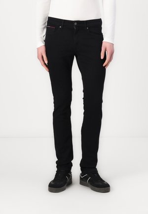 Person, die schmale schwarze Jeans, schwarze Sneakers und ein weißes Langarmshirt trägt, steht vor einem weißen Hintergrund.