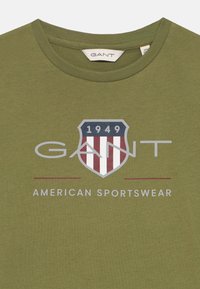 Olivgrünes Baumwoll-T-Shirt mit einem Schilddesign, roten und weißen Streifen, silberner Schrift "GANT" und "AMERICAN SPORTSWEAR" darunter.