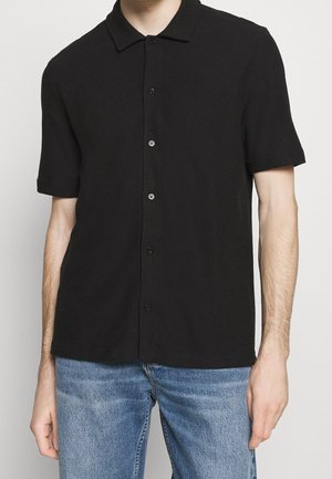 Homme portant une chemise noire à manches courtes avec des boutons et un jean bleu, debout devant un fond clair uni.
