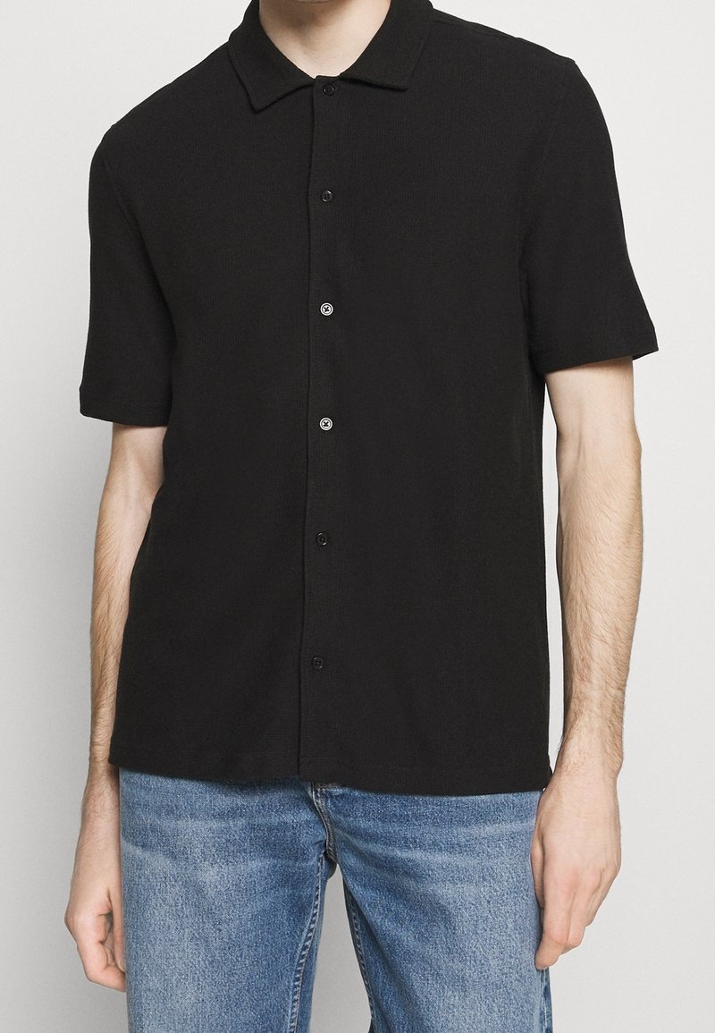 Homme portant une chemise noire à manches courtes avec des boutons et un jean bleu, debout devant un fond clair uni.