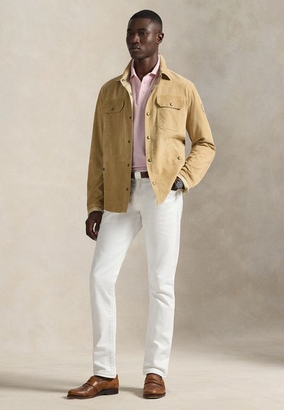 Άνδρας που φοράει μπεζ σακάκι πάνω από ροζ πόλο, λευκό παντελόνι, καφέ δερμάτινα μοκasin, όρθιος με το ένα χέρι στην τσέπη μπροστά σε ουδέτερο φόντο.