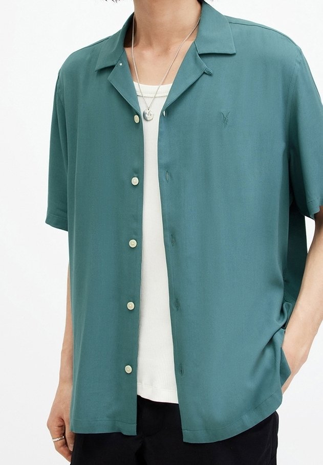 Kortarmig overhemd in teal stof, voorzien van een knoopsluiting aan de voorkant, een campkraag en subtiel logodetail. Draagt over een wit tanktopje.