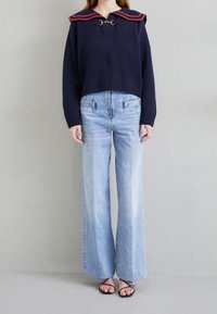 Marinblå kort tröja med röd-randig krage och metallbeslag, kombinerad med ljusblå vida jeans med framfickor och dragkedjor.