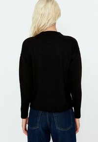 Henkilö, jolla on pitkät vaaleat hiukset, pukeutuneena mustaan neulepuseroon ja sinisiin farkkuihin, nähtynä takaa yksinkertaista taustaa vasten.