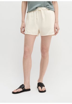 Osoba nosząca kremowe szorty ze sznurkiem i czarne sandały z odkrytymi palcami stojąca na jednolitym tle.