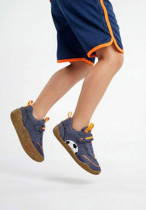 Kinderbeine in der Sprungmitte, tragen blaue und orangefarbene Shorts und blaue Sneakers mit orangen Akzenten und einem Fußballdesign an der Seite.