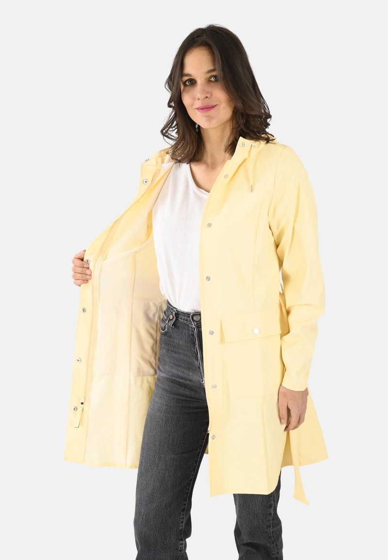 Imperméable jaune avec capuche, boutons-pression, poches et texture lisse ; porté par-dessus une chemise blanche et associé à un jean sombre.