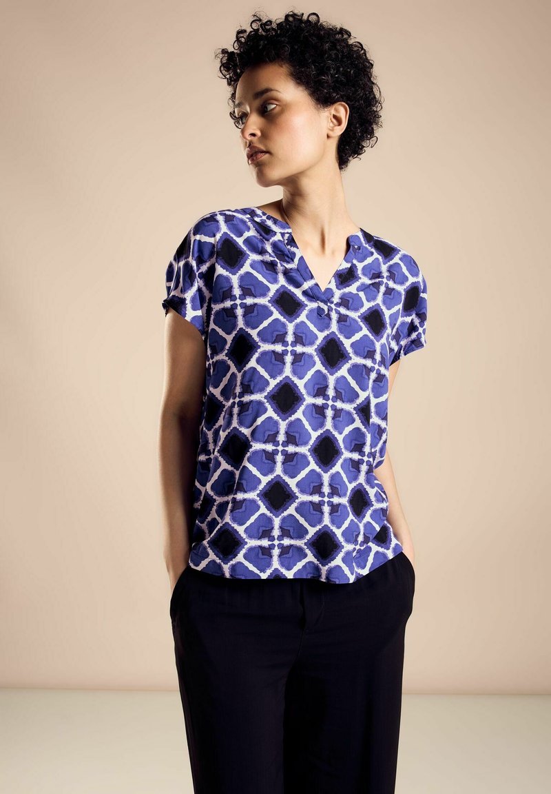 Short-sleeve blouse featuring a blue and white geometric pattern, with a v-neck and relaxed fit, paired with black trousers.