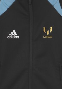 Black athletic zip jacket with light blue accents, featuring white Adidas logo and gold "MESSi" emblem on the chest. Soft fabric texture.