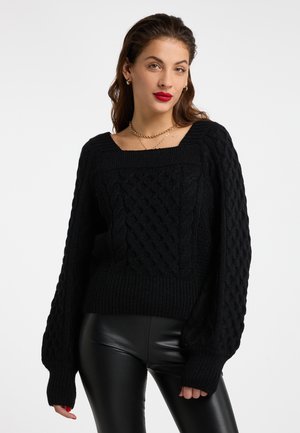 Frau mit schwarzem Zopfmusterpullover mit Ballonärmeln und schwarzer Lederhose, dazu goldene Halsketten und Creolen.