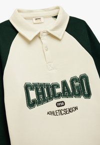 Cremefarvet polo-shirt med mørkegrønne raglanærmer. Har broderet "CHICAGO 1938" tekst og "ATHLETIC SEASON" i brun. Blødt stof.