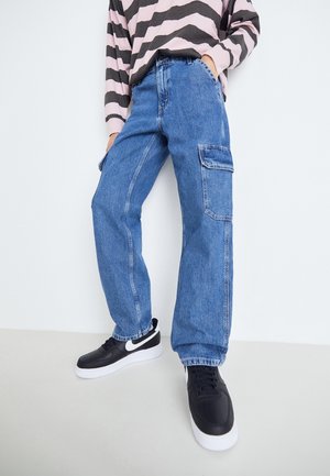 Person iført blå denim cargo jeans, sorte og hvide sneakers og en langærmet, stribet skjorte med den ene hånd i lommen.