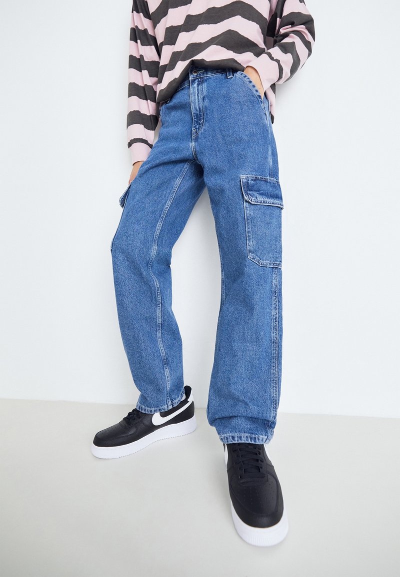 Personne portant un jean cargo en denim bleu, des baskets noires et blanches, et un t-shirt rayé à manches longues, une main dans la poche.