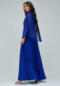 Vestido largo de color azul real con falda plisada, mangas de capa transparentes y sandalias de tacón alto plateadas. Tejido suave con una textura agradable.