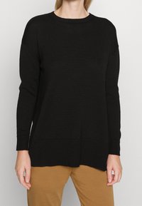 Femme portant un pull noir en tricot à manches longues avec un col rond et un pantalon marron, vue des épaules jusqu'aux cuisses supérieures.