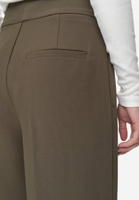 Bruine nauwsluitende broek met een achterzak, een gladde stofstructuur en een gestroomlijnd ontwerp. Hand dicht bij de taille geplaatst.