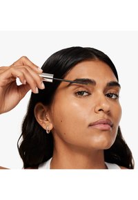 Una scovolino per mascara trasparente viene applicato su sopracciglia nere e folte. L'utente ha capelli neri lisci e pelle chiara, indossando gioielli minimi e una maglietta bianca.