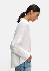 White button-up shirt with long sleeves and a loose fit, made from lightweight fabric. Paired with high-waisted blue denim jeans.
