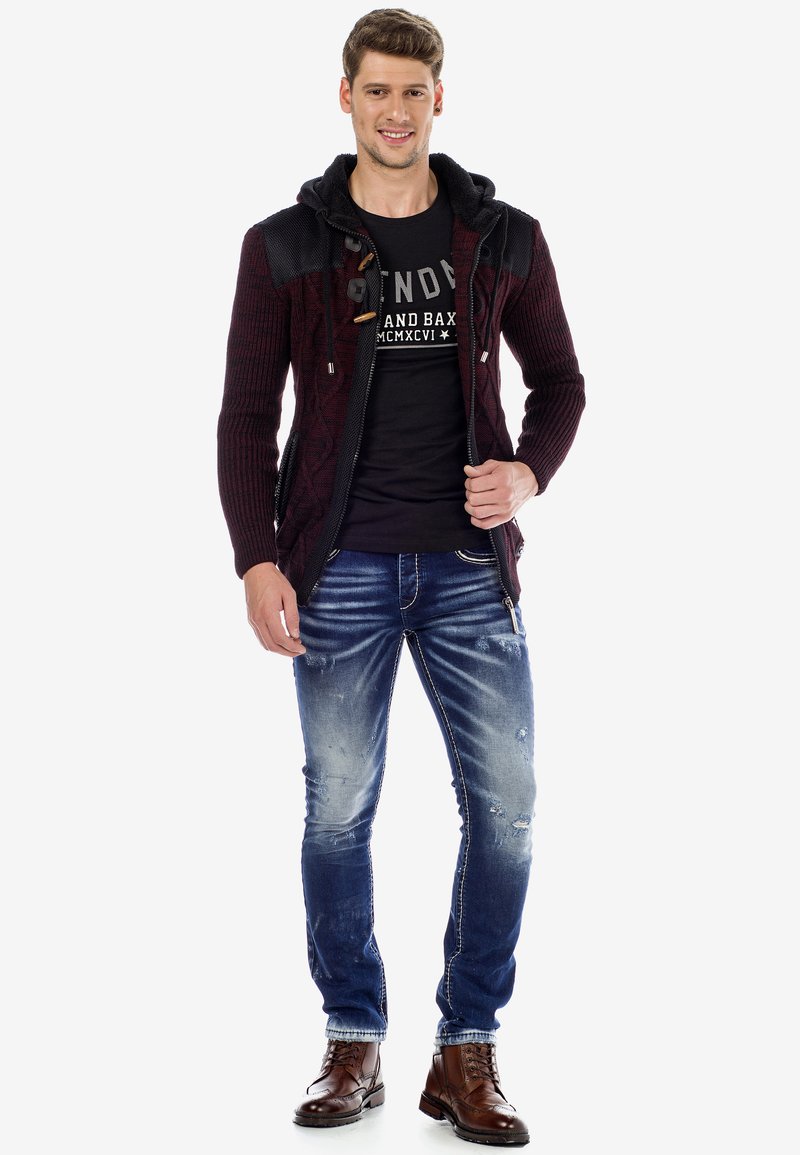 Haut de survêtement en maille bordeaux avec fermeture éclair, accents noirs, associé à un t-shirt graphique noir, un jean bleu déchiré et des bottes en cuir marron.