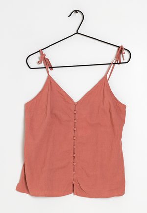 Blusa sin mangas color óxido con botones delanteros cubiertos de tela y tirantes ajustables con lazo colgando de una percha de metal negro.