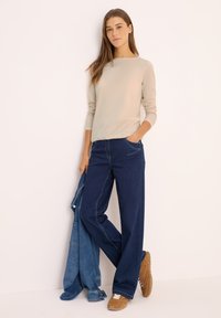 Beige long-sleeve top paired with wide-leg dark denim jeans. The model holds a light blue jacket, wearing brown sneakers with white laces.