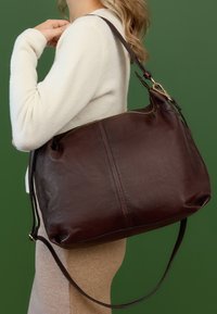 Borsa a spalla in pelle marrone con una trama liscia, chiusura con zip superiore e tracolla regolabile. Presenta un corpo arrotondato e cuciture in contrasto.