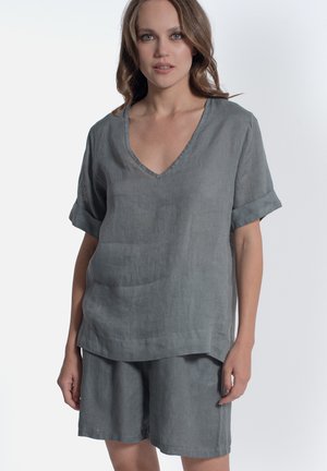Donna che indossa un top grigio di lino a maniche corte con scollo a V e pantaloncini abbinati, in piedi davanti a uno sfondo chiaro e uniforme.