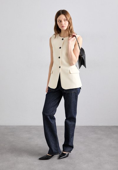 Gilet sans manches ivoire avec des boutons noirs, doté de poches latérales, associé à un jean droit bleu foncé et des chaussures noires à bout pointu.