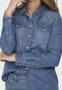 Frau trägt ein blaues Denim-Hemd mit weißen Druckknöpfen und Brusttaschen, Arme verschränkt und Hand berührt das Gesicht, kombiniert mit blauen Jeans.