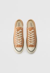 Baskets en toile couleur beige avec lacets blancs, embout en caoutchouc, bordure de semelle noire et détails de couture. Tige texturée avec des accents de couleur contrastante.
