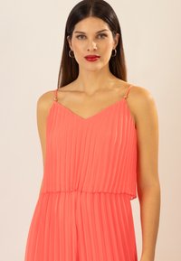 Vestido de coral con plisados y tirantes finos que presenta detalles dorados, escote en V y un diseño fluido que añade textura y movimiento.