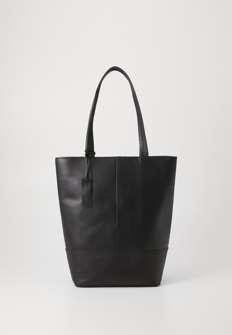 Sac cabas en cuir noir avec deux longues anses et une petite étiquette rectangulaire attachée, debout sur un fond blanc uni.