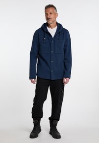 Veste polaire bleu marine avec capuche, dotée de deux poches avant, de boutons pression, et pantalons cargo noirs avec poches latérales. Bottes noires à lacets.