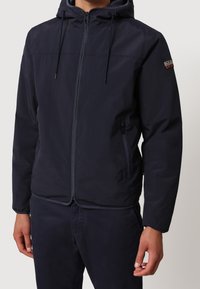 Giacca leggera blu navy con cappuccio, zip frontale e cordini regolabili. Dotata di tasche laterali e di un patch con logo sulla manica.