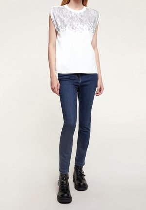 Haut blanc sans manches avec détail texturé en sequins argentés sur les épaules, associé à un jean slim bleu foncé et des bottines noires à lacets.