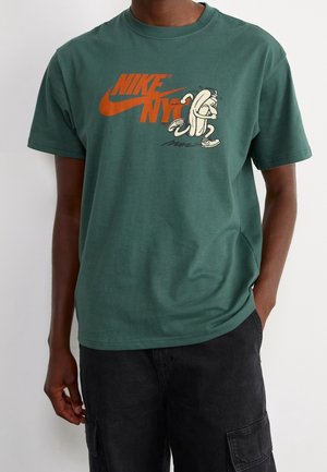 Mand iført en grøn Nike T-shirt med orange "NIKE NYC" tekst og et tegneserieagtigt løbende bananmotiv, parret med sorte cargo shorts.