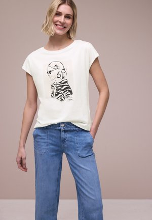 Uśmiechnięta blondynka, ubrana w białą koszulkę z czarno-białą ilustracją kobiety w okularach przeciwsłonecznych, w połączeniu z jasnoniebieskimi dżinsami, neutralne tło.