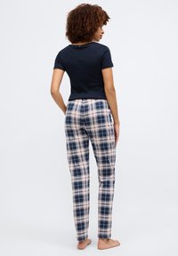 Femme aux cheveux bouclés debout pieds nus, de dos, portant un t-shirt bleu marine à manches courtes et un pantalon à carreaux rose et bleu marine sur fond blanc.