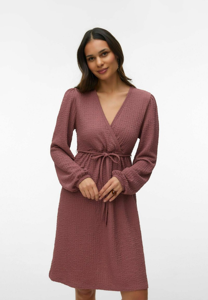 Femme aux longs cheveux noirs portant une robe portefeuille mauve texturée à manches longues, nouée à la taille, debout devant un fond clair uni.