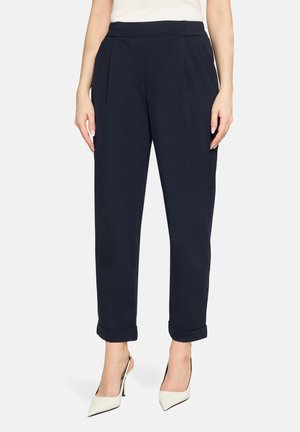 Pantalon bleu marine fuselé avec plis, tissu doux, taille élastique et revers roulés, associé à des chaussures blanches à talons hauts.