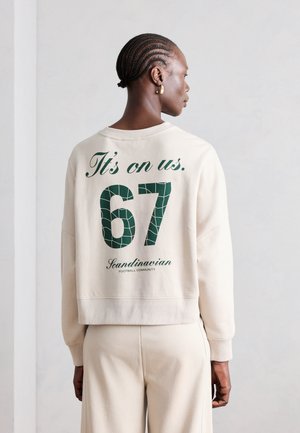 Oseba z pletenicami, oblečena v kremasto jopa in hlače, s hrbtom proti kameri; na jopi piše "It's on us. 67 Scandinavian Football Community."