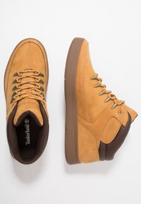 Helle Wildlederschuhe im High-Top-Stil mit braunen Ledermustern, metallischen Ösen und einer Gummisohle. Verfügt über einen gepolsterten Kragen für zusätzlichen Komfort.