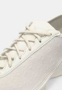 Κοντινή λήψη ενός λευκού δερμάτινου αθλητικού παπουτσιού με υφασμάτινη πλευρική πάνελ και μπεζ κορδόνια σταυρωμένα πάνω στη γλώσσα.