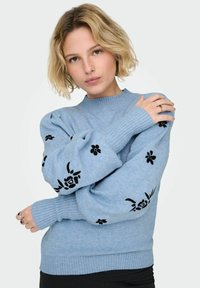 Světle modrý pletený svetr s nabíranými rukávy, zdobený černou květinovou výšivkou na rukávech a žebrovanými manžetami a lemem. Měkká textura.